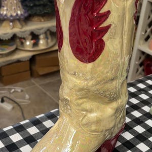 May include: A decorative ceramic cowboy boot vase. The boot is a pearlescent cream color with a red wing design and a red heel and sole. The vase is on a black and white checkered surface.