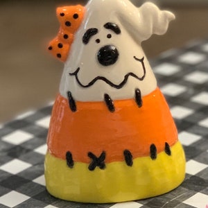 May include: A white ghost with a black and orange candy corn body and a black and orange polka dot bow. The ghost has a black nose and black eyes.