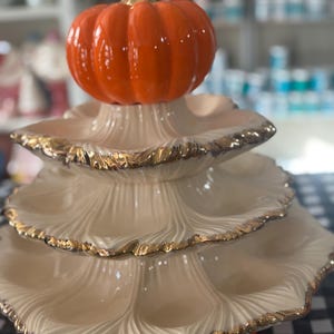 May include: A tiered serving dish with a pumpkin topper. The dish has three cream-colored layers with gold trim and an orange base. The pumpkin is orange with a gold stem. Perfect for fall decor.