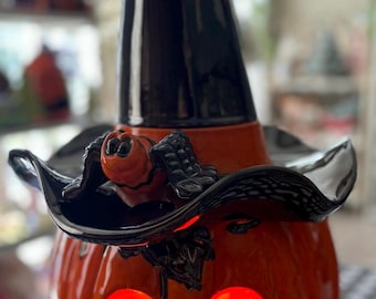 May include: A ceramic jack-o'-lantern figurine featuring a glossy orange pumpkin base with illuminated cut-out eyes and a mouth. A tall, black, pointed witch's hat sits atop the pumpkin, adorned with a small, sculpted black spider on the brim.