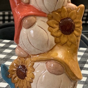 May include: A stack of three ceramic gnomes, each wearing a sunflower hat. The gnomes are painted in shades of brown, beige, and blue. The bottom gnome is wearing a blue hat and has a sunflower on its chest.