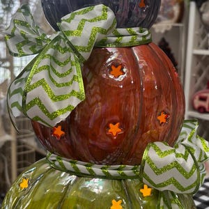 May include: A stacked pumpkin decoration featuring three glossy ceramic pumpkins in purple, orange, and green. Each pumpkin has star-shaped cutouts and is adorned with a white and green glittery chevron ribbon tied in bows between the layers.