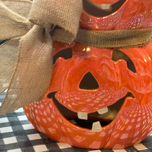 May include: Two orange ceramic pumpkins with a burlap bow. The pumpkins have a carved jack-o'-lantern face with a wide smile and teeth.