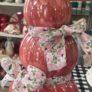 May include: A stacked pumpkin decoration made of three glossy, coral-pink glass sections with a metallic gold stem. The structure is wrapped with a white ribbon featuring a repeating pattern of pink pumpkins and green leaves, tied into two bows.