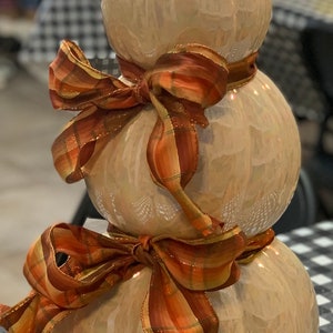 May include: A stack of three white ceramic pumpkins with a brown and orange plaid ribbon tied around them. The pumpkins are decorated with a pearlescent finish.