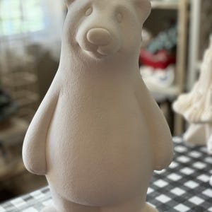 May include: A white ceramic bear figurine with a simple, rounded design. The bear has a small, rounded nose and small, black eyes. The bear is standing on a black and white checkered surface.
