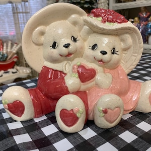 May include: A pair of ceramic teddy bears holding hearts, one wearing a red dress and the other a pink dress. The bears are sitting on a black and white checkered surface.