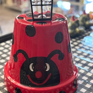 May include: A red ladybug-themed flower pot with black polka dots and a smiling ladybug face. A small black lantern sits atop the pot. The pot is on a black and white checkered surface.