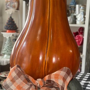 May include: A tall, glossy, orange ceramic gourd-shaped pumpkin sits atop a smaller, matte, sage green pumpkin. A decorative fabric bow featuring a brown, orange, and white plaid pattern is tied around the center where the two pumpkins meet.