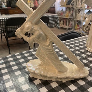 May include: A white ceramic figurine of a person carrying a cross. The figurine is standing on a base that looks like a rock. The figurine is made of a shiny, iridescent material.