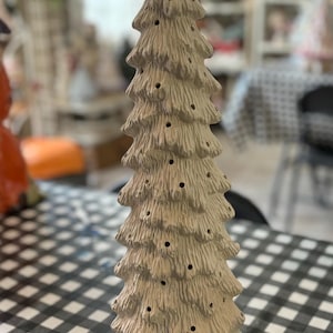 May include: A ceramic Christmas tree with a textured, off-white finish. The tree has multiple tiers with small, evenly spaced holes for lights. It sits on a round base and is displayed on a black and white checkered tablecloth.