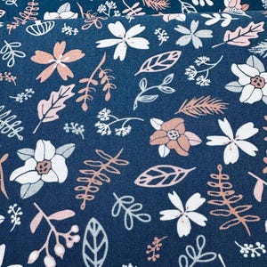 May include: Close-up of a navy blue fabric with a floral pattern. The design features white and peach-colored flowers, along with various leaves and sprigs in white, peach, and gray. The overall aesthetic is a delicate, botanical theme.