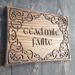 May include: Wooden sign with carved Celtic knot border and the words "céad míle fáilte" in a stylized font. The sign is made of light brown wood with a dark brown border and text.