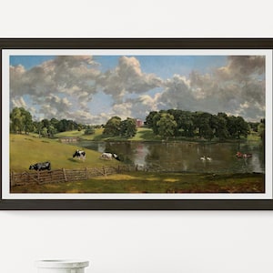 May include: Framed landscape art print featuring a pastoral scene with cows grazing in a green field near a lake. The artwork depicts a cloudy sky, trees, and a red building in the distance. The frame is dark brown.