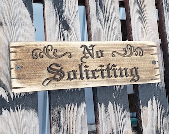 May include: Wooden sign with the words "No Soliciting" carved in a decorative font. The sign is rectangular, with a light brown finish and dark brown text. The sign is mounted with screws.