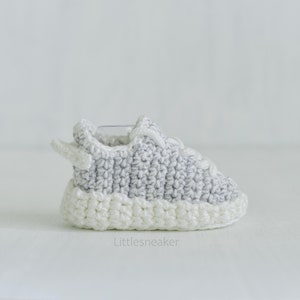 May include: A pair of gray and white crocheted baby shoes. The shoes have a white sole and a gray upper with a white lace.