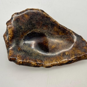 May include: A handcrafted, organic-shaped ceramic dish with a glossy, mottled brown and tan glaze. The dish has a shallow, irregular form, perfect for holding small items. The interior features a darker, pooling effect, adding depth and visual interest.