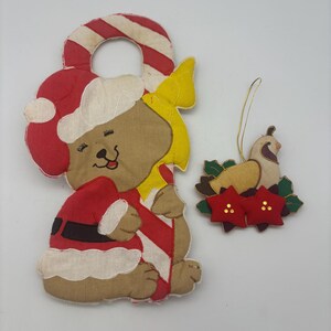 May include: A fabric door hanger featuring a tan bear wearing a Santa hat and suit, holding a red and white candy cane, alongside a smaller fabric ornament of a bird perched on green leaves with two red flowers and gold accents.