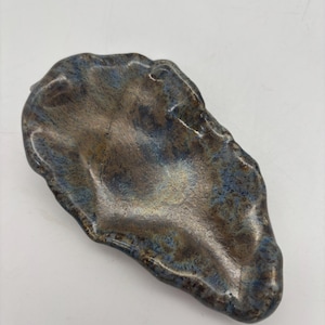 May include: A ceramic oyster-shaped dish with a metallic finish. The dish has a textured surface with a blend of blue, brown, and gold hues. The irregular shape and color variations give it a unique, handcrafted appearance. Ideal for serving small appetizers or as a decorative piece.