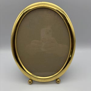 May include: An oval-shaped picture frame with a polished gold-tone metal finish. The frame features a smooth, rounded border and is supported by two small, spherical ball feet at the base, allowing it to stand upright on a flat surface.