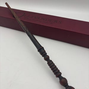 May include: A dark brown wooden wand with a ribbed handle and a tapered shaft featuring carved spiral details. The wand is positioned in front of a long, rectangular, burgundy-colored box with the text Ollivanders Makers of Fine Wands since 382 BC.
