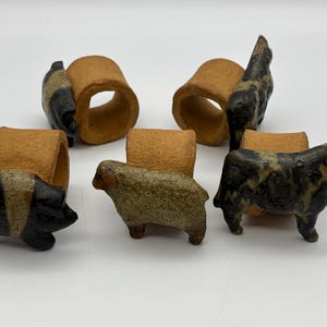 May include: Set of five ceramic napkin rings. Each ring has a tan-colored cylindrical base with a small animal figurine attached. The animals include a pig, sheep, cow, and two other animals with black and tan markings.