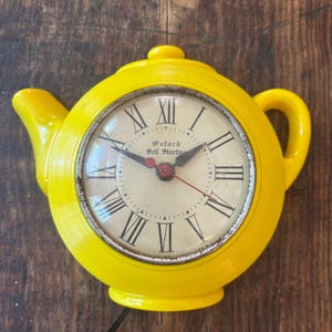 May include: A bright yellow teapot-shaped wall clock with a round body, a spout, and a handle. The clock face is off-white with black Roman numerals, black hour and minute hands, and a red second hand. The text Oxford Self Starting is printed on the face.