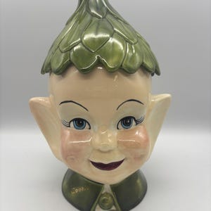 May include: A ceramic cookie jar shaped like an elf head with a green leaf-patterned hat lid. The elf has large pointed ears, blue eyes, rosy cheeks, and a dark red mouth. The word Cookie is written in gold script on the green collar base.