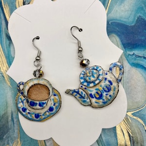 Puede incluir: Un par de pendientes colgantes con una tetera azul y dorada y una taza de té azul y dorada. Los pendientes están hechos de metal y tienen un acabado plateado.