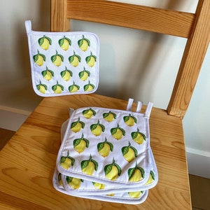 May include: White square pot holders with a repeating pattern of yellow lemons with green leaves. The pot holders have a white border and a small white loop for hanging. The pot holders are stacked on a wooden chair.