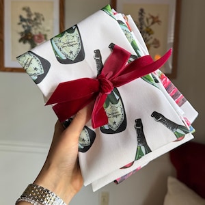 May include: White cloth napkins with a champagne bottle pattern, tied with a red velvet ribbon. The napkins feature a repeating design of green, black, and gold champagne bottles. The napkins are folded and stacked.