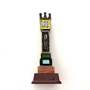 May include: A black and gold miniature replica of the Gastown Steam Clock in Vancouver, Canada. The clock has a white face with black hands and Roman numerals. The base is made of brown wood.