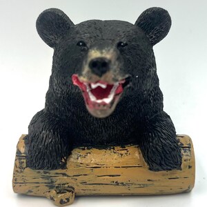Peut inclure: Figurine d'ours noir avec la bouche ouverte, assis sur une bûche brune. L'ours a une fourrure noire et un nez brun.
