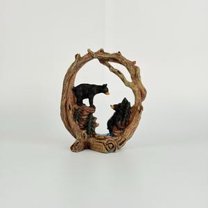 Peut inclure: Une figurine décorative représentant deux ours noirs debout sur un terrain rocheux à l'intérieur d'un cadre circulaire en forme de tronc d'arbre brun texturé. La scène comprend de petits pins verts et un élément d'eau bleu à la base des ours, le tout rendu dans une sculpture détaillée en trois dimensions.