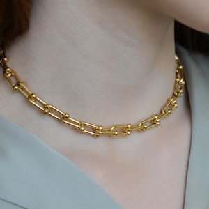 May include: A gold-toned chain necklace featuring elongated rectangular links connected by small, polished spherical beads at each joint. The necklace rests around the neck, showcasing a uniform metallic finish and a structured, repeating link design.