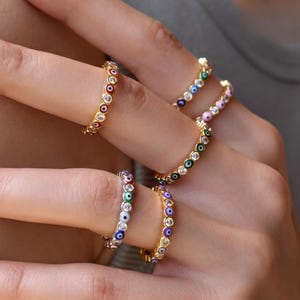 May include: A collection of gold-toned rings featuring a continuous band of small, circular evil eye charms in various colors including red, green, blue, purple, and pink, interspersed with clear, sparkling stones set in a metallic finish.
