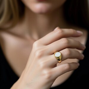 May include: A gold signet ring with a white oval center stone. The ring is worn on a finger, showcasing its polished gold band and the smooth, light-colored stone. The ring's design is simple and elegant.