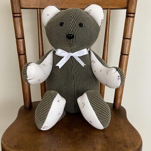 May include: A stuffed teddy bear crafted from olive green corduroy fabric with white fabric accents featuring small pink floral patterns on the ears, arms, and feet. The bear has black button eyes and a black nose, finished with a white satin ribbon.