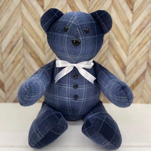 May include: A stuffed teddy bear crafted from blue and grey plaid fabric. The bear features dark round button eyes, a black button nose, and three black buttons down the center of its torso. A white satin ribbon is tied in a bow around its neck.