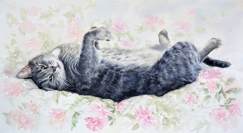 May include: A watercolor painting of a grey tabby cat lying on its back with its paws curled inward. The cat has dark grey stripes, white whiskers, and green eyes. It is positioned against a soft, light background featuring pale pink flowers.