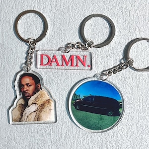 Könnte beinhalten: Drei Schlüsselanhänger mit silbernen Ringen. Einer zeigt ein Porträt eines Mannes in einem Pelzmantel. Ein anderer hat das Wort "DAMN." in Rot. Der dritte Schlüsselanhänger zeigt ein Bild eines schwarzen Vans.