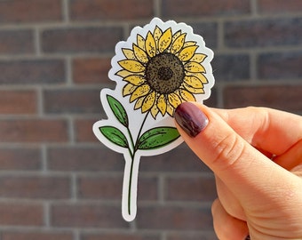 May include: A sticker featuring a hand-drawn illustration of a sunflower with yellow petals and a brown center. The sunflower has a green stem and two leaves.