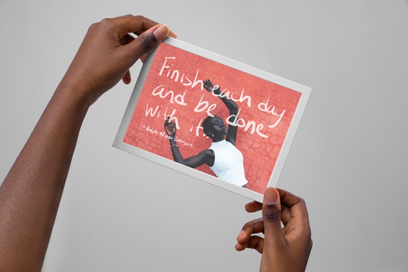Op de afbeelding: Een ingelijst print met een citaat dat luidt: "Maak elke dag af en wees er klaar mee." - Robin Williams. De print toont een foto van een persoon met een donkere huidskleur die voor een rode muur staat.
