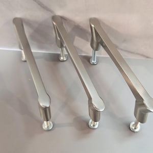 May include: Three brushed nickel cabinet pulls with a curved, elongated design. Each handle features two cylindrical mounting posts with visible silver-toned screws extending from the base, providing a sleek and uniform metallic appearance.