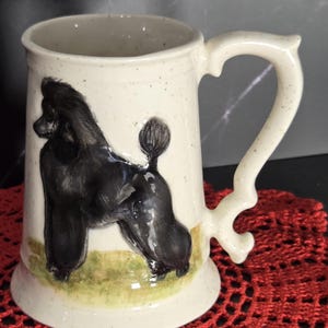 May include: A cream-colored ceramic mug with a speckled finish and a decorative handle. The mug features a raised, hand-painted black poodle standing on a patch of green grass. The design is centered on the front of the cylindrical vessel.