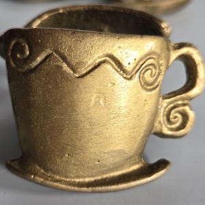 May include: A small, gold-toned metal cup-shaped object with a flat base and a handle. The exterior surface features a raised, embossed zigzag line pattern that runs horizontally across the upper portion, ending in spiral details near the handle.