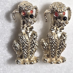May include: A pair of gold-toned poodle figurines. Each poodle has red rhinestone eyes, a black nose, and a bow tie. The poodles are standing upright with textured fur detail. These vintage-style figurines are likely brooches or earrings.