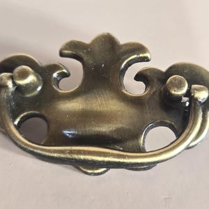 May include: A brass-toned metal drawer pull featuring a decorative backplate with a central fleur-de-lis shape and two side wings. A curved, swinging bail handle is attached to the backplate, allowing for a grip to open a drawer or cabinet door.