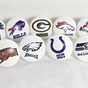 May include: A collection of eleven white, egg-shaped decorative items, each featuring a different NFL team logo. Logos include the Ravens, Lions, Bills, Packers, Broncos, Buccaneers, Vikings, Bengals, Eagles, Colts, and Seahawks.
