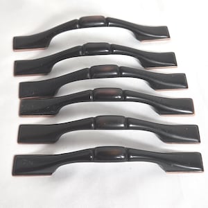 May include: Six dark brown cabinet handles with a curved design. Each handle has a rectangular center section and rectangular ends. The handles are arranged horizontally against a white background. The handles are made of a dark brown material with a copper-colored edge.